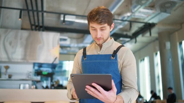 Male cafe worker using tablet device in restaurant interior. Man wearing apron checking digital information on screen. Hospitality management. Reviewing orders while standing near bar counter in cafe.