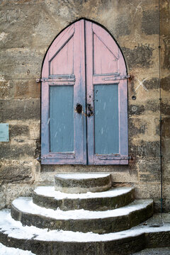 View of a weathered, pastel-painted arched door stands against a rough stone wall, framed by snow-dusted steps leading up to it, Rothenburg ob der Tauber, Bavaria, Germany.