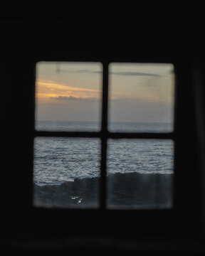 View of the sea's dark waves crashing against the shore under a muted sunset sky, all framed by the panes of a weathered window, Clachtoll Beach, Sutherland, Scotland.