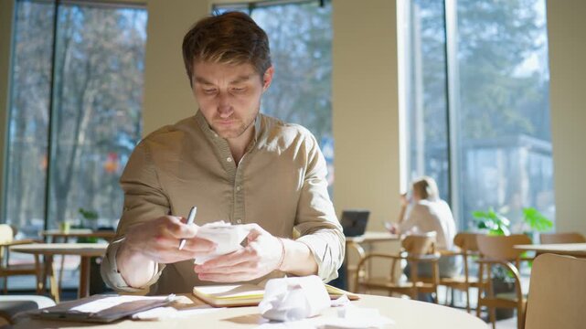 Male worker sitting at table with stack of receipts. Holding papers and documents. Looking down and analyzing expenses. Notebook placed on surface. Organizing records. Cafe interior with large windows
