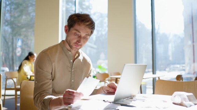 Man sitting at table with laptop and receipts. Holding papers and touching head. Looking stressed and thinking. Checking numbers and reviewing documents. Paper checks scattered on desk.
