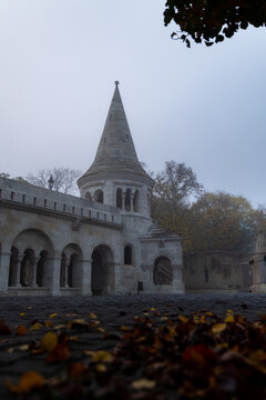 View of the Fisherman's Bastion's striking white stone turrets pierce through the dense fog, with fallen leaves scattered on the ground, Budapest, Hungary.