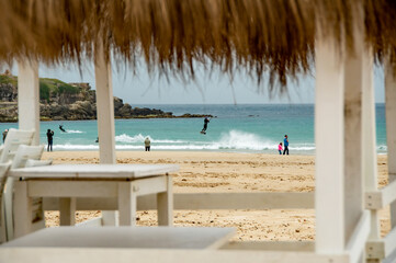 Kite surfers in the waves on a windy day. Spectators watch on the sandy beach, Bright colourful kites fill the cloudy sky. Turquoise water. copy space.
