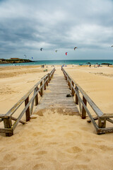 Boardwalk, wooden path leading to the beach. Kite surfers in the waves on a windy day. Bright colourful kites fill the cloudy sky. Turquoise water and sandy beach copy space.