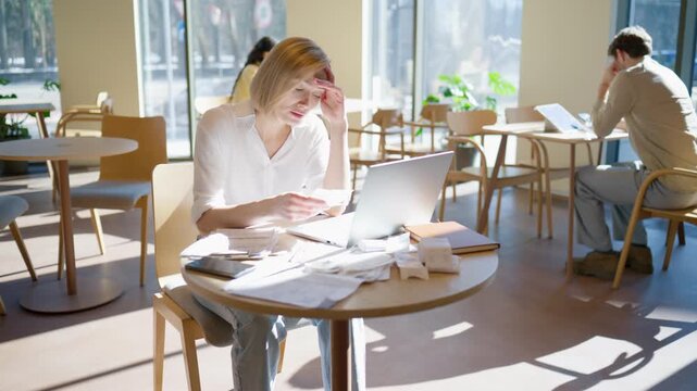 Woman sitting at table with laptop and receipts. Covering mouth and checking papers. Fixing payment mistake and reviewing calculations. Documents scattered around. Cafe interior with daylight.
