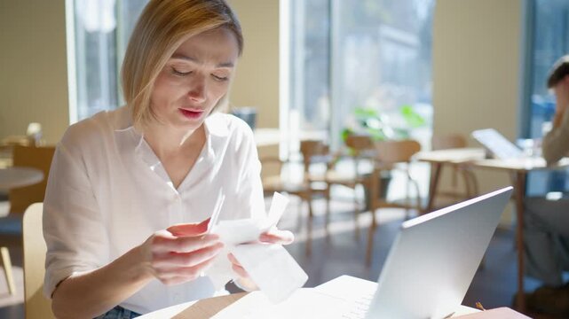 Caucasian woman sitting at table and holding many receipts. Looking confused and stressed. Checking numbers and reviewing documents. Laptop placed on desk. Managing payments and workload.