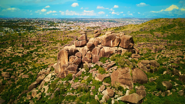 Aerial view of colossal boulders and verdant vegetation dominate the foreground, contrasting with the distant cityscape under a bright sky, Jos, Plateau, Nigeria.