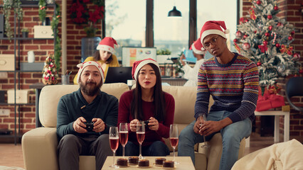 Naklejka premium Diverse coworkers playing video games on console during christmas party celebration in office. Using controller to play gaming competition with colleagues, celebrating winter holiday. Tripod shot.