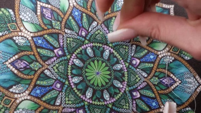 Close up of female hands sewing beads on decorative canvas with intricate pattern. Handmade craft concept showing patience, creativity, traditional needlework and textile art.