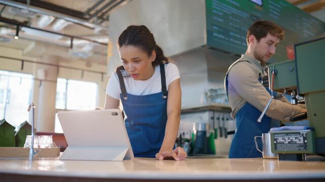 Barista woman standing at counter and using tablet device. Leaning forward and reading screen carefully. Male coworker preparing coffee at machine. Wearing aprons. Handling tasks simultaneously.