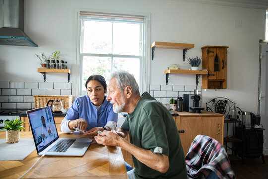 Nurse assisting elderly man with telemedicine consultation in home kitchen