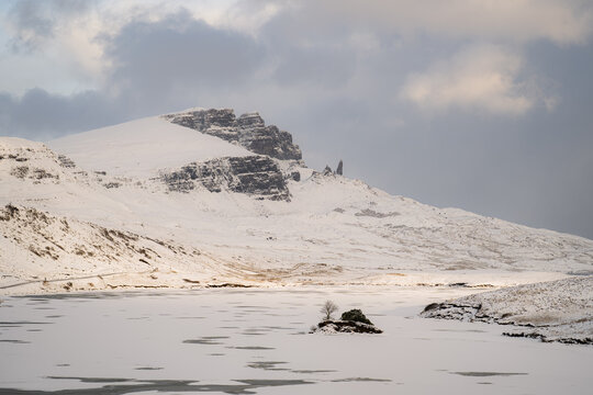 View of a snow-blanketed landscape with rugged peaks piercing the sky, an islet in a frozen Loch Fada adds a touch of serene beauty, Old Man of Storr, Isle of Skye, Scotland.