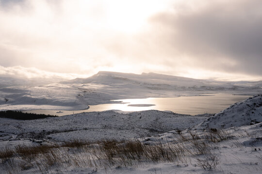 View of a serene landscape where snow blankets the rolling hills of Rasaay Island, under a soft, diffused light, reflecting on the Sound of Rasaay, Isle of Skye, Scotland.