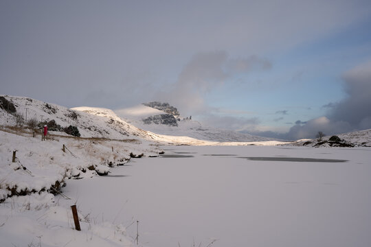 View of a snow-covered landscape with a frozen Loch Fada and rocky mountains in the background under a cloudy sky, casting shadows on the ground, Old Man of Storr, Isle of Skye, Scotland.