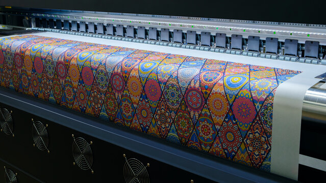 digital printer rolls out colorful, intricate patterned fabric. Vibrant geometric designs cover the material with rich hues. Pattern features mandala-inspired motifs, blending tradition