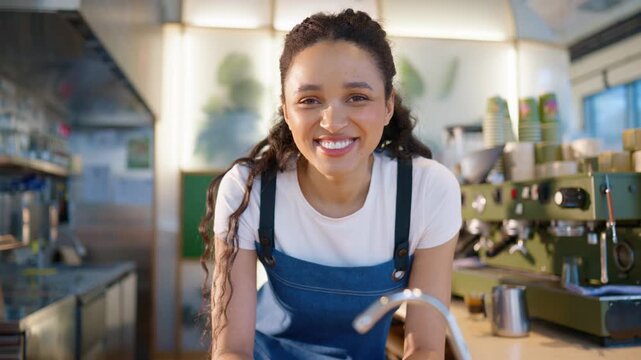 Female barista leaning on counter and looking at camera. Young woman smiling while pausing from work activity. Wearing apron and standing near coffee machine. Waiting for next order inside cafe.