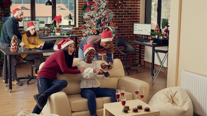 Fototapeta premium Cheerful colleagues playing video games at christmas party in office, feeling happy and enjoying winter holiday celebration. Using controllers on console to play gaming competition at festivity.