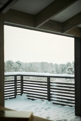 Fototapeta premium Snow covered balcony railing overlooking winter forest landscape with trees in background, outdoor scene showing untouched snow accumulation on wooden structure and ground, vertical