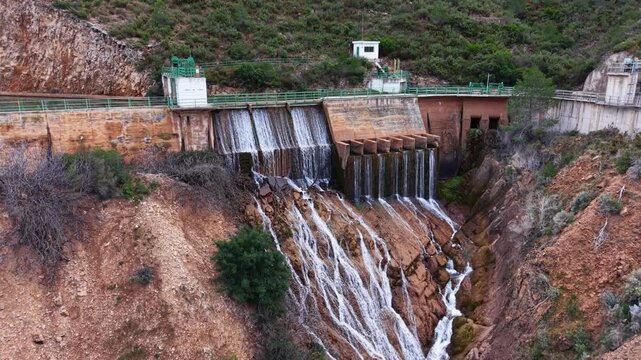 Aerial view of a concrete dam spillway releasing water over stepped weirs into a rocky channel, showcasing hydraulic engineering, erosion patterns, and water management in a rugged landscape