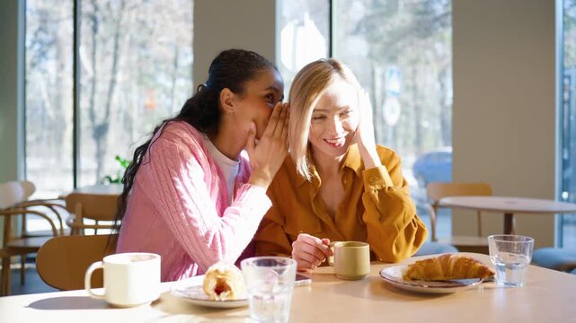 Two female friends sitting at cafe table with breakfast. Woman whispering secret while covering mouth with hand. Friend smiling and listening with curiosity. Drinking coffee and sharing private gossip