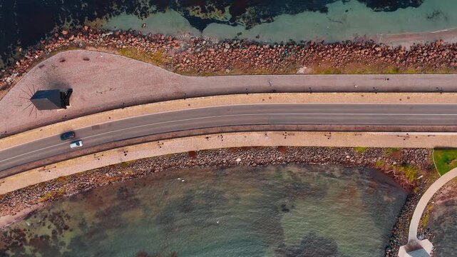 Top down aerial view of coastal road and windmill. Aerial view of a paved road on an island isthmus with a wooden windmill, rocks, and sea water.