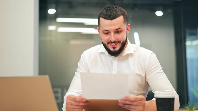 Cheerful professional male executive with beard in white shirt sitting at office desk and smiling at documents. Successful businessman or lawyer happy about positive results or profit in office