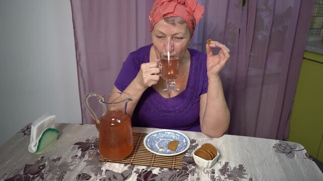 Elderly woman drinking compote from a glass at home. Pour a drink from a carafe. Eating cookies