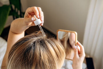 Fototapeta premium Woman applying hair serum with dropper to scalp while lying on a bed, focusing on hair care and beauty routine