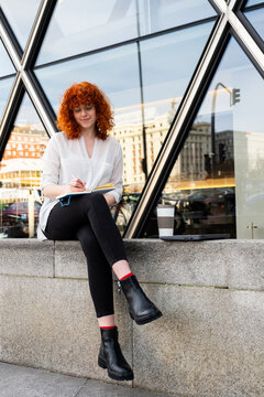 Redhead writing in notebook outside urban building with coffee