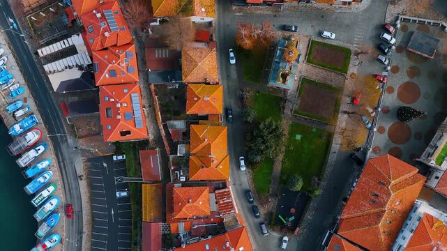 Aerial view of city square and fishing harbor. Overhead shot of a town square with green space, red roofs, and blue boats docked along the quay.