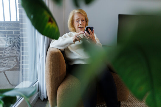 Senior sitting in armchair using phone in cozy living room with plants
