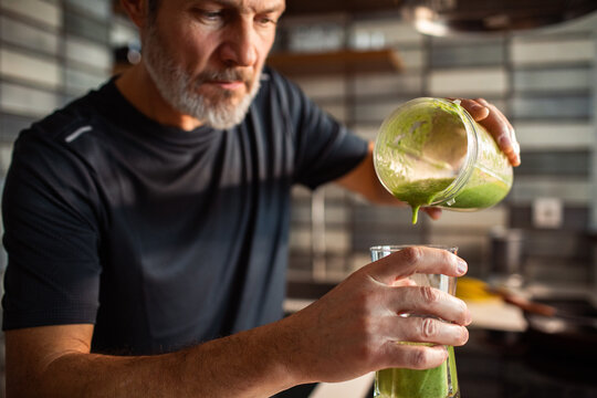 Mature man pouring green smoothie into glass in home kitchen