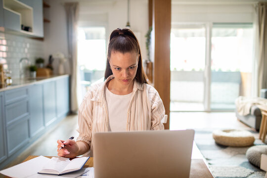 Focused young woman working on laptop at home kitchen