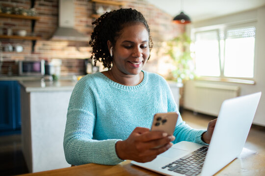 Smiling woman using smartphone and laptop at home kitchen