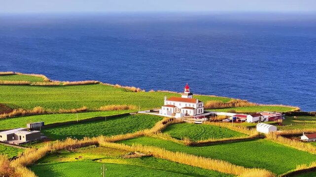 Aerial view of Farol da Ponta do Arnel lighthouse perched on a lush green cliff above the deep blue Atlantic Ocean in the Azores, Portugal.