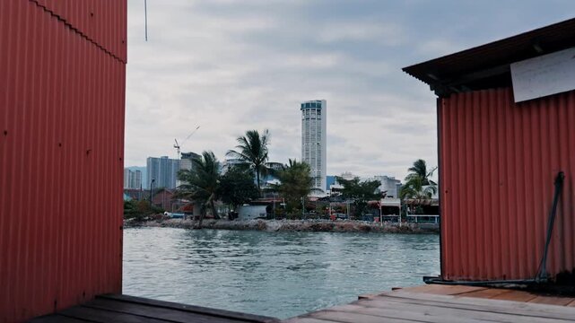 A view across the water toward the modern George Town skyline and the iconic Komtar Tower, framed by the rustic orange corrugated metal walls of a traditional clan jetty.