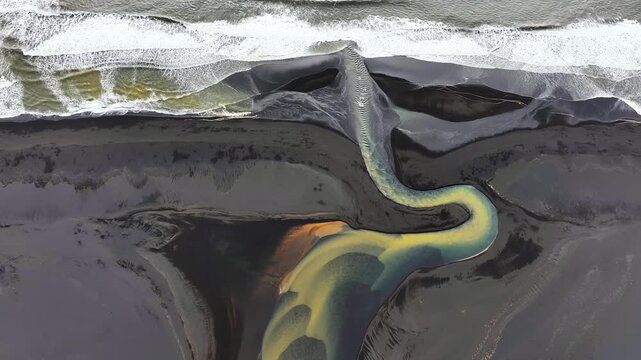 Aerial top-down view of powerful Atlantic waves colliding with glacial river on Iceland&rsquo;s volcanic black sand coast, forming dramatic natural patterns where ocean water meets river flow.