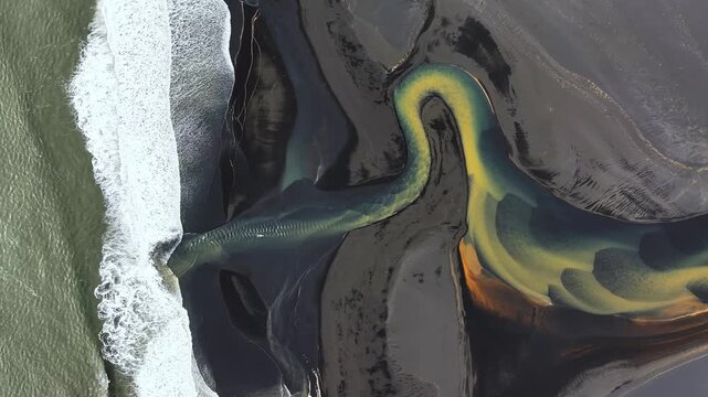 Aerial view of colorful river meeting Atlantic Ocean along Iceland&rsquo;s black sand coast, striking patterns where glacial water flows into the sea. Top down.