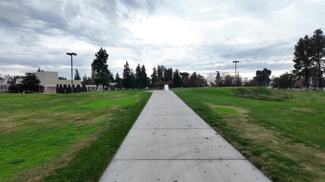 First-person perspective moving forward along a concrete sidewalk in a public park, surrounded by green grass fields and trees under a cloudy sky, representing a journey forward