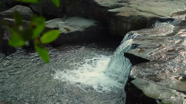 Small waterfall spilling over flat rock ledges into a round pool, capturing fresh flowing water, dark stone texture, and peaceful creek scenery in a natural setting.