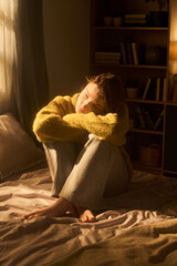 Young woman sitting on bed hugging knees, gazing thoughtfully toward window, sunlight illuminating face, bookshelf and home interior in background © Mediaphotos