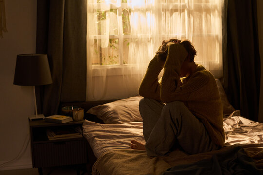Young woman sitting on bed holding head with both hands, knees drawn to chest, appearing stressed or overwhelmed in softly lit bedroom near window with curtains