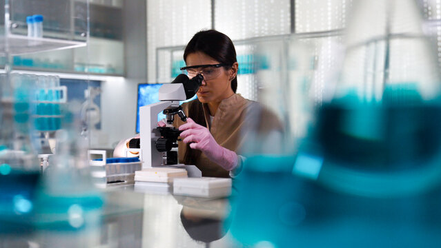 Female scientist in protective gear working in a modern laboratory to test human DNA samples. Looking into the microscope. Selective focus
