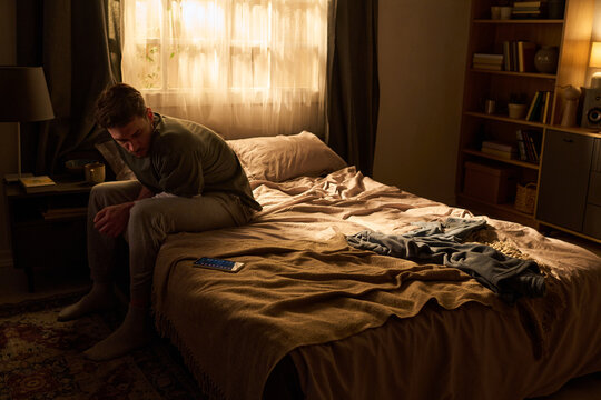 Young man sitting on edge of bed looking down, resting elbows on knees in dimly lit bedroom, smartphone lying on blanket, casual clothing scattered beside him