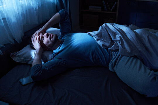 Adult man lying in bed holding head with both hands appearing distressed, smartphone resting beside pillow, bedroom illuminated by soft blue light from window
