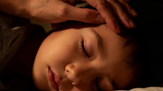 Reaching caregiver hand settling on sleeping child in grey shirt at bedside, checking for fever