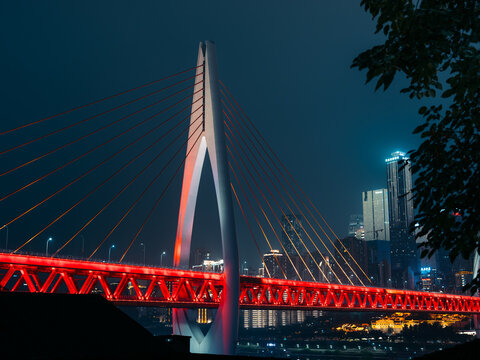 View of a modern cable-stayed bridge illuminated with vibrant red lights piercing the dark night sky, contrasting with the city skyline, Chongqing, Chongqing, China.