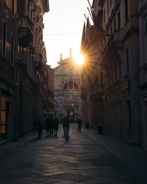 View of the sunburst illuminating the architecture and pedestrian traffic along a narrow street, creating a warm glow, Venice, Veneto, Italy.