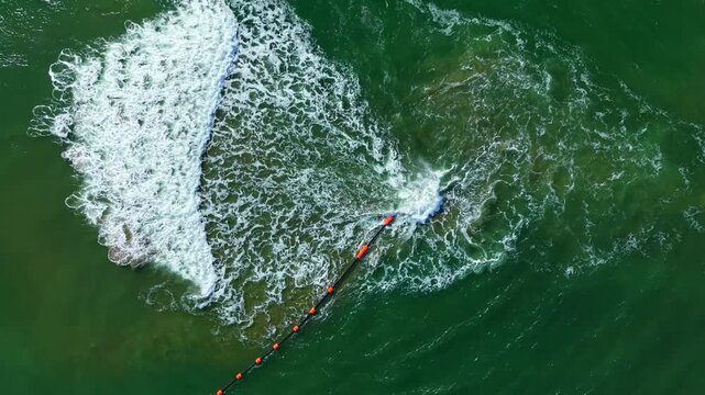 A surfboard rides on the waves near the breakwater while orange buoys mark the area. The sun shines on the water, and the surf creates foam, showing the energy of the sea.