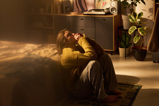 Young woman sitting on floor beside bed holding hands near face appearing thoughtful illuminated by warm indoor light with houseplants and furniture in background